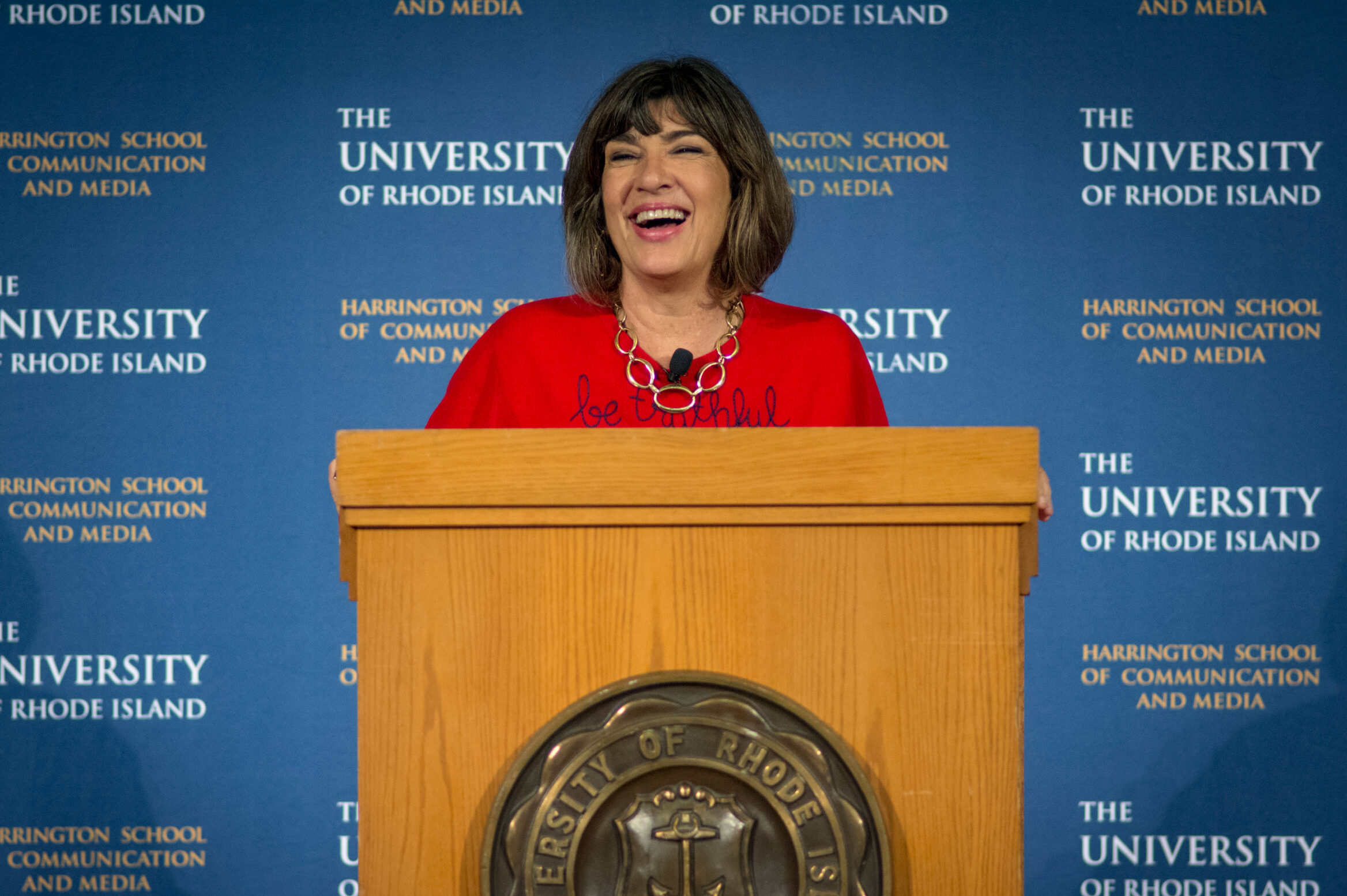 克里斯蒂安·阿曼普尔站在演讲台上笑着，背景是罗德岛大学的标志。